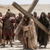 Devine Bible Guide - Jesus Christ Carrying the Cross - www.javascapeai.com A man depicted as Jesus carries a large wooden cross through a desert while Roman soldiers and onlookers surround him.