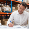Business professional working on paperwork beside a laptop in a warehouse with stacked boxes and storage racks in the background.