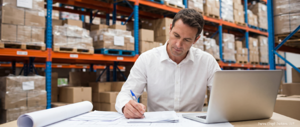 Business professional working on paperwork beside a laptop in a warehouse with stacked boxes and storage racks in the background.