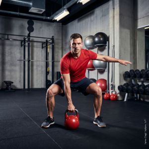 Athlete in a red shirt performing a kettlebell exercise in a modern gym with dumbbells, stability balls, and strength equipment in the background.