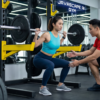 Coach guiding an athlete performing a barbell squat in a modern gym with weight plates, racks, and a JAVASCAPE AI Gym sign in the background.