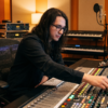 Music producer adjusting a large studio mixing console with audio tracks displayed on computer monitors, a microphone, keyboards, acoustic panels, and rack-mounted studio equipment.