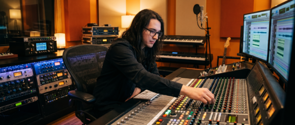 Music producer adjusting a large studio mixing console with audio tracks displayed on computer monitors, a microphone, keyboards, acoustic panels, and rack-mounted studio equipment.