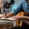 Songwriter Pro - javascapeai.com Songwriter with guitar writing in a notebook beside a laptop, studio microphone, desk lamp, and crumpled paper.