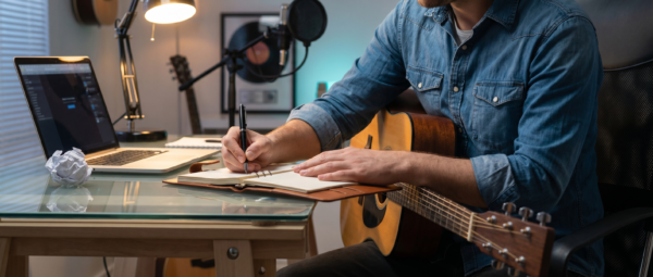 Songwriter Pro - javascapeai.com Songwriter with guitar writing in a notebook beside a laptop, studio microphone, desk lamp, and crumpled paper.