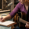 Songwriter Pro - javascapeai.com Songwriter with guitar writing in a notebook beside a laptop, studio microphone, and crumpled paper.