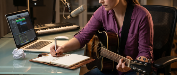 Songwriter Pro - javascapeai.com Songwriter with guitar writing in a notebook beside a laptop, studio microphone, and crumpled paper.