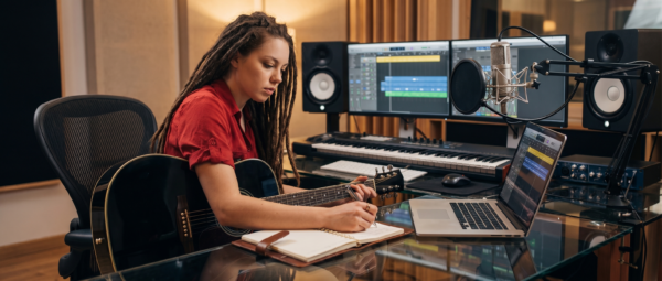 Songwriter Pro - javascapeai.com Songwriter with guitar writing in a notebook beside a laptop, studio microphone, monitors, and keyboard in a music studio.