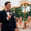 Man in a tuxedo holding a microphone while giving a wedding speech as seated guests smile and laugh under warm reception lights.