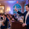 Speechwriter Assistant — Pro - javascapeai.com Man in a suit raising a champagne glass while giving a wedding reception toast from a podium as seated guests watch.