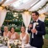Man in a tuxedo holding a microphone while giving a wedding reception speech as the bride and guests smile under soft draped lighting.