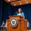 Speechwriter Assistant — Pro - javascapeai.com - Product Page 4 Woman speaking at a wooden podium on a formal stage with seated attendees, blue curtains, microphones, and warm auditorium lighting.