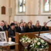 Speechwriter Assistant — Pro - javascapeai.com - Product Page 73 Woman speaking at a lectern during a church memorial service with seated attendees, floral arrangements, a candle, a portrait, and a grand piano nearby.