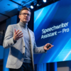 Speechwriter Assistant — Pro - javascapeai.com Man presenting on stage beside a large blue screen displaying “Speechwriter Assistant — Pro” during a professional speaking event.