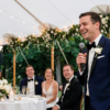 Groom giving a speech with a microphone at an outdoor wedding reception while seated guests laugh beneath a decorated marquee.