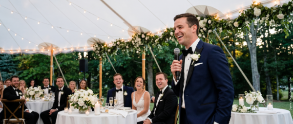 Groom giving a speech with a microphone at an outdoor wedding reception while seated guests laugh beneath a decorated marquee.
