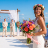 Bride holding a colourful bouquet at a beach wedding ceremony with seated guests, a floral arch, and the ocean in the background.