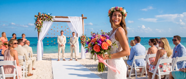 Bride holding a colourful bouquet at a beach wedding ceremony with seated guests, a floral arch, and the ocean in the background.