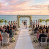Beach wedding ceremony at sunset with a floral arch, seated guests, palm trees, and the ocean in the background.