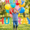 Young child jumping with colorful balloons beneath a large “Hurray” sign in a