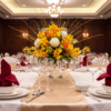 Formal banquet table set with white linens, burgundy napkins, glassware, gold accents, and a yellow floral centrepiece in an elegant event room.