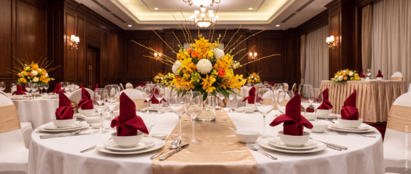 Formal banquet table set with white linens, burgundy napkins, glassware, gold accents, and a yellow floral centrepiece in an elegant event room.