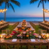 Long outdoor dining tables decorated with tropical flowers, fruit, candles, and hanging lights beside the ocean at sunset.