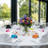 Round event table set with white linens, folded napkins, glassware, colorful place cards, candles, and a vibrant floral centrepiece.