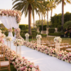 Outdoor wedding ceremony with a white floral arch, pink and ivory roses, gold chairs, a flower-lined aisle, palm trees, and a live band.